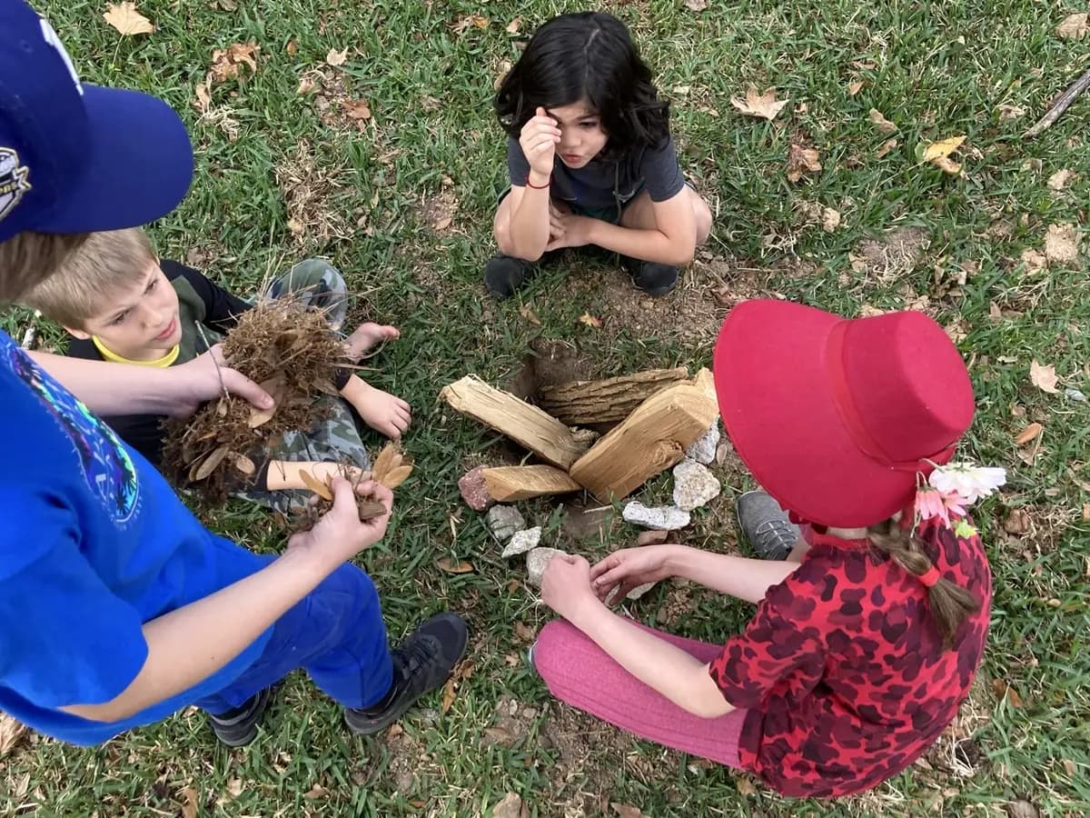 Learners gathered around a small fire pit, holding dry leaves and split wood.